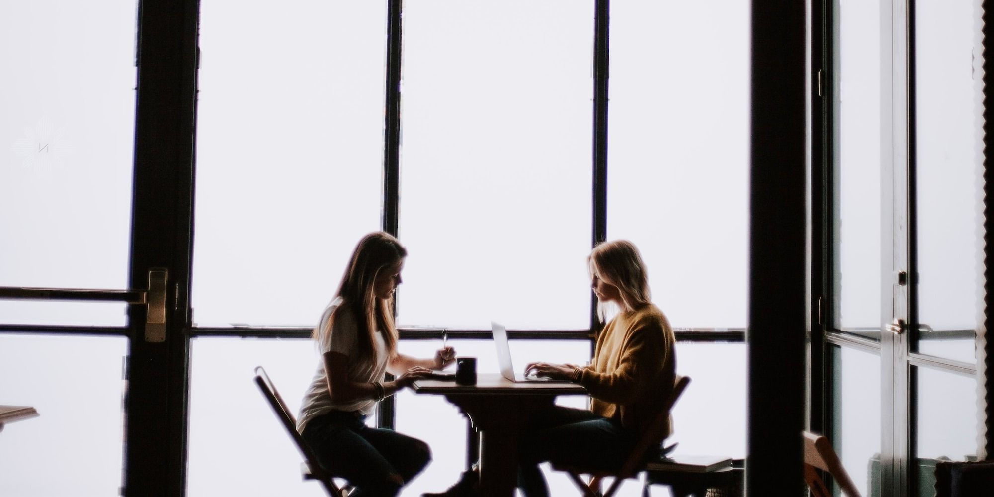 photo of two women facing each other sitting in front of table near glass wall